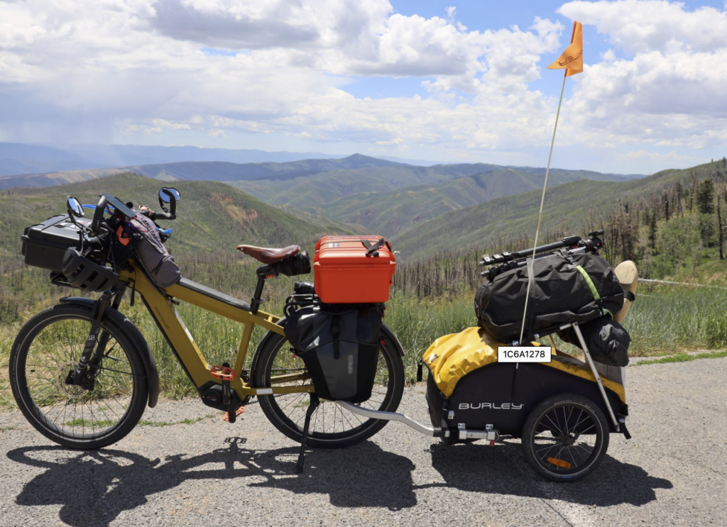 Touring bicycle loaded with front and rear panniers and an orange dry box, parked on a mountain road with rolling hills and a partly cloudy sky in the background.
