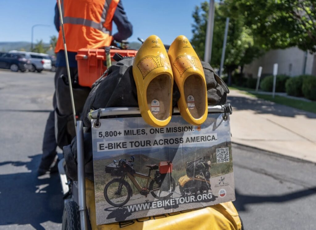 Yellow clogs perched on a bicycle rack over a poster that reads '5,800+ miles, one mission: E-bike tour across America'.